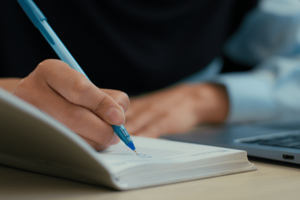 Person documenting car accident settlements information while writing notes with blue pen next to laptop