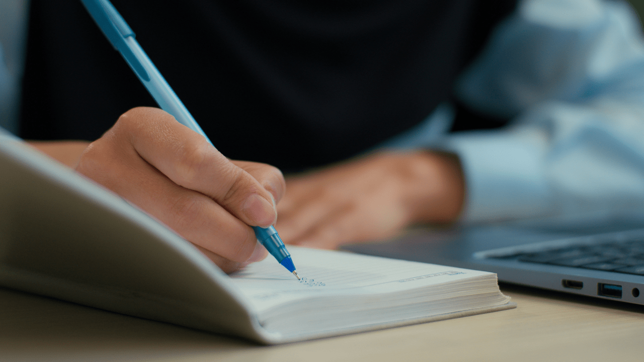 Person documenting car accident settlements information while writing notes with blue pen next to laptop