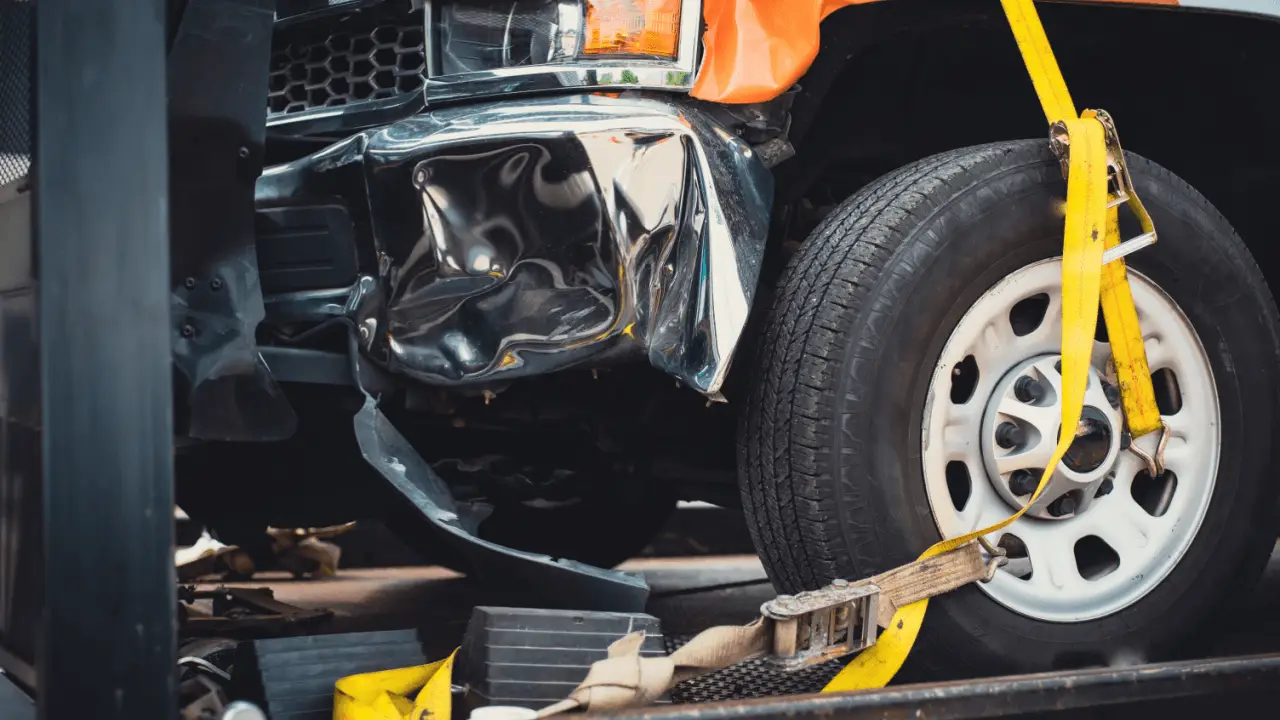 Truck accident liability scene with severely damaged vehicle showing crushed front fender and hood secured by yellow straps on tow truck